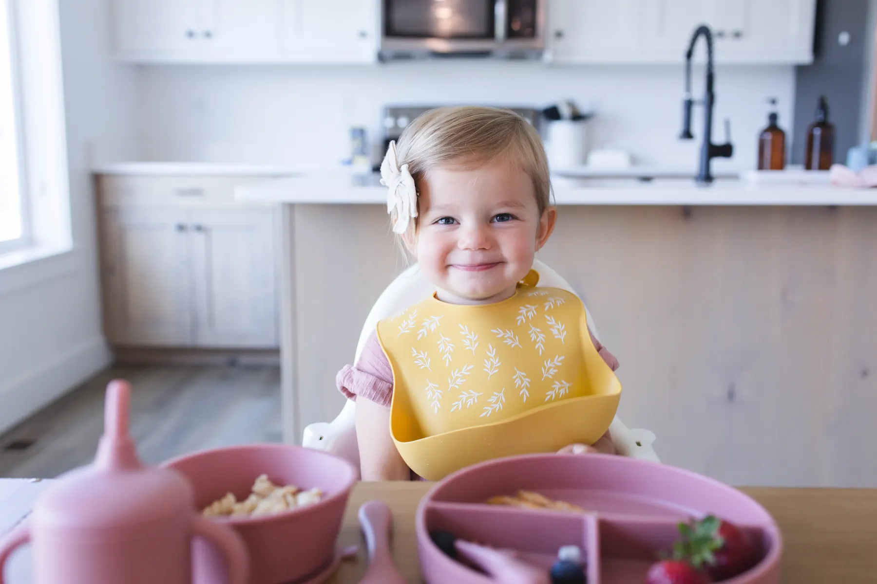 A smiling baby at a high chair wearing a yellow KAYA silicone bib during a self-feeding meal